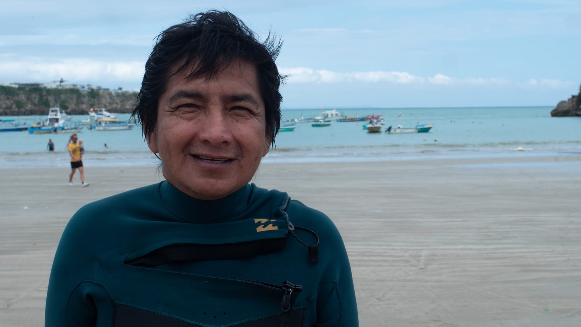 Ambrosio Yagual wearing a diving suit with fishing boats behind him in Ayangue, Ecuador